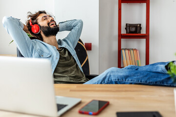 Smiling young adult hipster man relaxing at workplace. Happy freelancer male entrepreneur listening...