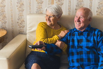 Laughing aged caucasian couple, man and woman a comedy show on watching tv while sitting on a beige...