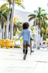 Back view of the walking girl with afro hairstyle, carrying skateboard in hand. Summer vibes. Hobby concept