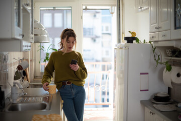 Woman standing in the kitchen and drinking cup of tea while using smartphone