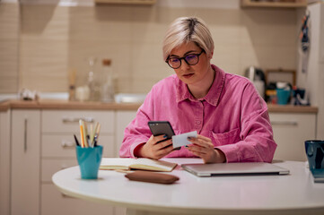 Woman with a blond short hair holding credit card and using smartphone at home