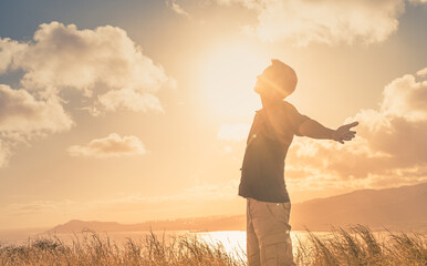 It's a beautiful life! Young man with arms up to the sunlight standing in a meadow field. - obrazy, fototapety, plakaty