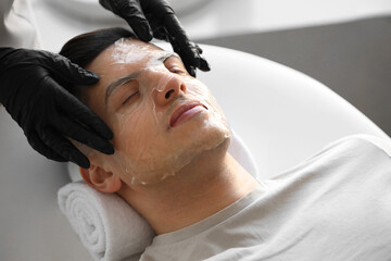 Fototapeta na wymiar Cosmetologist applying gel mask onto young man's face in salon, closeup