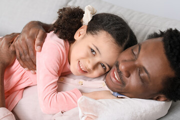 Happy African-American man and his little daughter lying on sofa at home