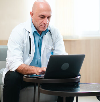 Male doctor is using notebook computer in hospital living room. Professional medical person in white coat and stethoscope sits relaxing looking at computer screen for diagnosis, healthcare information