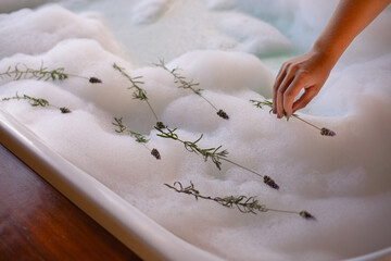 A woman's hand placing sprigs of lavender on top of the foam of a bathtub