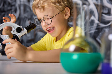 Young scientist of five years squinting carefully looks with one eye into eyepiece of microscope...