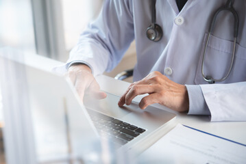 Doctor working on laptop computer with medical chart clipboard on table at doctor's office in...