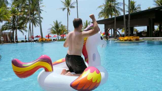 Funny man having fun, dancing on floating inflatable unicorn on tropical vacation in swimming pool in modern luxury resort. Summer holidays, sunny day, enjoying summer, sea view on background. 