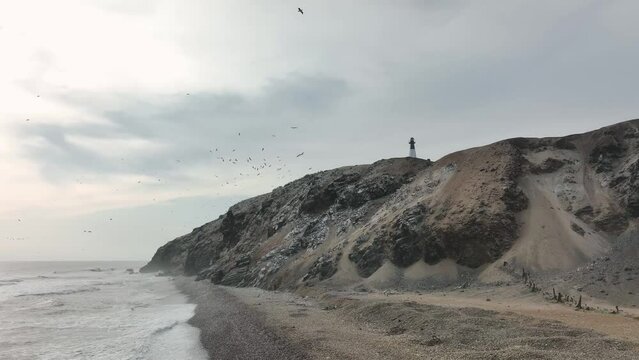 Aerial view of the breaking waves on the rocky coast with a lighthouse, birds and cliffs in the background. Media Luna beach, Puerto Eten, Lambayeque, Peru.