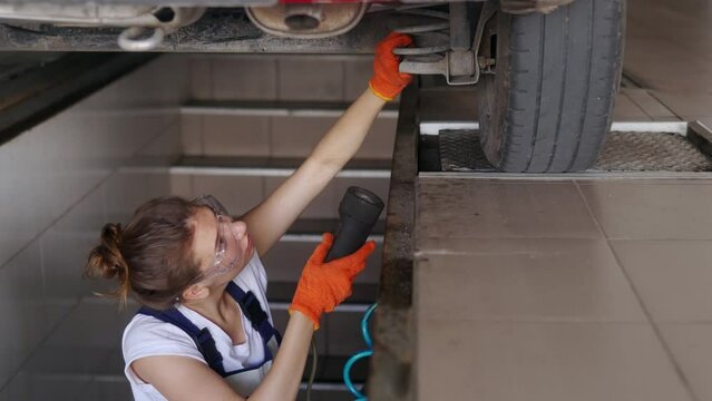 Female mechanic inspecting car suspension on hydraulic wheel play detector with remote control. Strong woman checks for wear in running gear and steering components. Service operator in overalls.