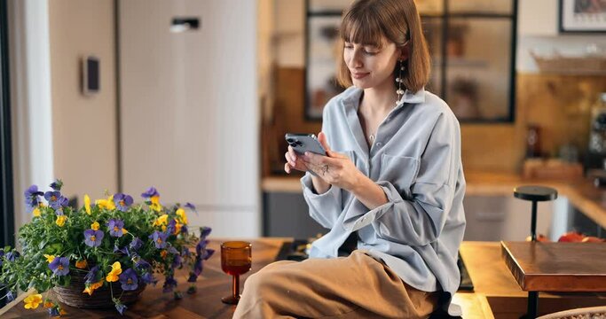 Young woman sits on a kitchen top and using phone, spending leisure time in beautiful modern house. Home coziness and household life concept