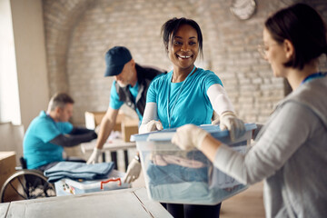 Happy African American volunteer passing box with packed clothes to her coworker at donation center.