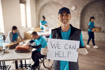 Happy mature man holding placard with 'we can help' message while volunteering at charity center.