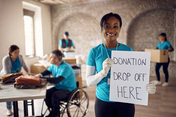 Happy African American woman holding placard with 'donation drop off here' while volunteering at...