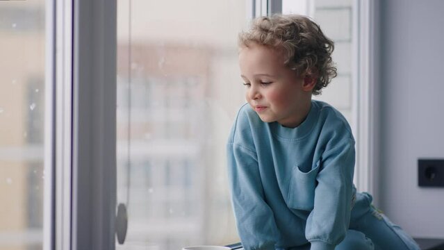 chubby slavic little boy with charming curly hair is sitting on windowsill in room in snowy day, looking out window
