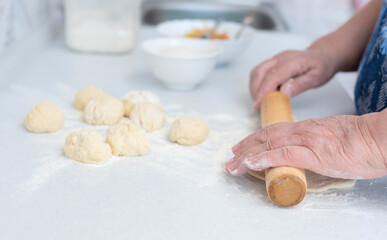 Senior woman hands rolling out the dough with a rolling pin on a white kitchen table with blurred grated apple and sugar on background. Selective focus. Process of making pies with apple filling