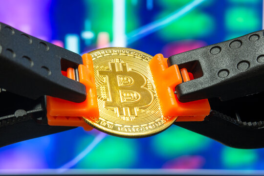 Representative bitcoin between plastic clamps in selective focus. Bitcoin on market chart background. Concept of blocking bitcoin, stopping its rise, crypto currency, stock market, digital money.