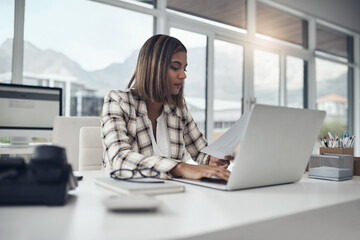 I need to go over all the business accounts today. Shot of an attractive young businesswoman sitting alone in her office and using her laptop while reading paperwork. - obrazy, fototapety, plakaty