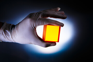 A hand in a white medical glove, in which there is a rectangular chemically manufactured glass for a laser. Bright light