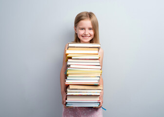 Primary school girl holding stack of books on a grey wall background
