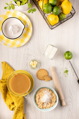 Lemon cake ingredients: lemons, limes, crushed cookies, butter and more laid out on a luminous kitchen wooden table
