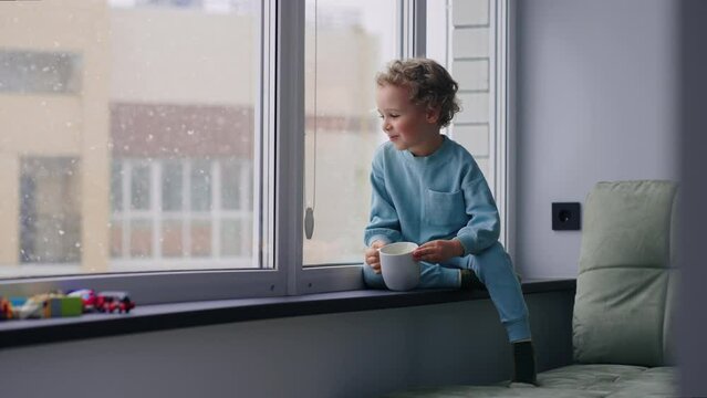 cute chubby boy in blue pajamas is watching first snow in window and drinking milk from cup in home