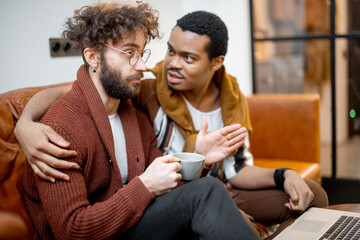 Gay couple with different nationality having close conversation while sitting on a couch at home. Concept of homosexual relations and lifestyle at home. Caucasian and hispanic man together indoors
