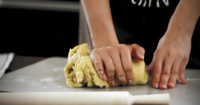 Close-up. A pretty brunette woman in a white T-shirt and a brown apron kneads dough for sweet pastries. Women's manicured hands are kneading shortbread dough on a silicone mat.