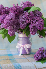 Vertical photo of a bouquet of flowers with lilac petals in a vase with a bow on a table with a checkered tablecloth. Copy space on the left