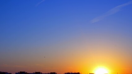 The setting sun over the roofs of city buildings in the distance is landing a passenger plane. Beautiful urban sunset. The blue sky is flooded with the yellow fire of the setting sun