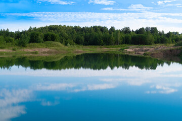 Fototapeta na wymiar Picturesque blue lake summer landscape for outdoors vacation. Blue sky and clouds reflection in clear pond water with lush greenery forest in the background.