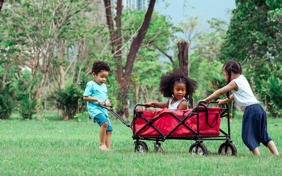 Mixed Race Little Cute Children Helping Each Other To Push And Drag A Red Cart While Playing In Outdoor Green Park. Education, Diversity, Unity And Teamwork Concept.