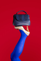 A blue foot in a beige shoe with high heels is raised up on a red background. Women's legs upside down with a blue handbag on high-heeled shoes.