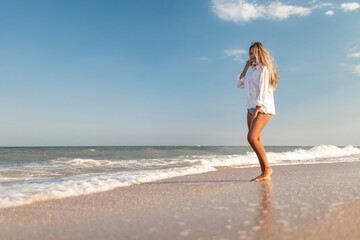 Fototapeta na wymiar A slender girl in a gentle blue swimsuit and shirt walks along the sandy beach near the blue sea