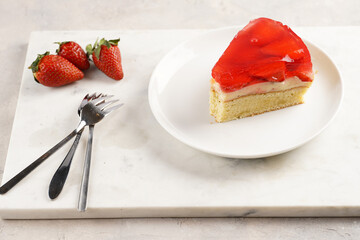 a Piece of a three-layered cake with vanilla pudding, strawberry jelly and fresh strawberries on a white marble background