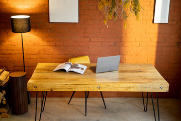 Stylish and cozy workplace with a wooden table and floor lamp on brick wall background at home. Laptop and magazine on the table top