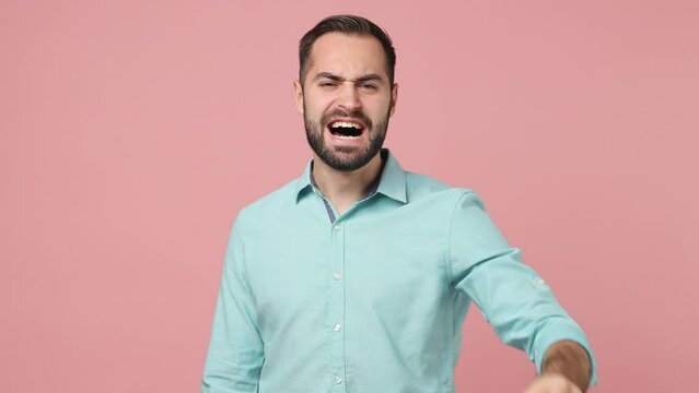 Displeased irritated sad angry mad young brunet man 20s wears blue shirt scream swear shout call out gesticulating with hands pointing finger on camera isolated on plain plain pink background studio