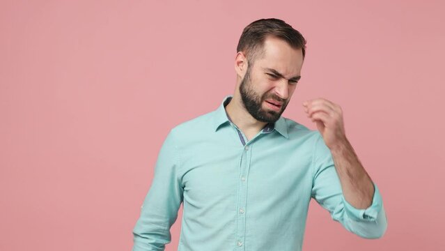 Irritated angry young brunet man 20s wears blue shirt looking aside feeling bad ugly smell waving hand closing nose isolated on plain plain pink background studio. People emotions lifestyle concept