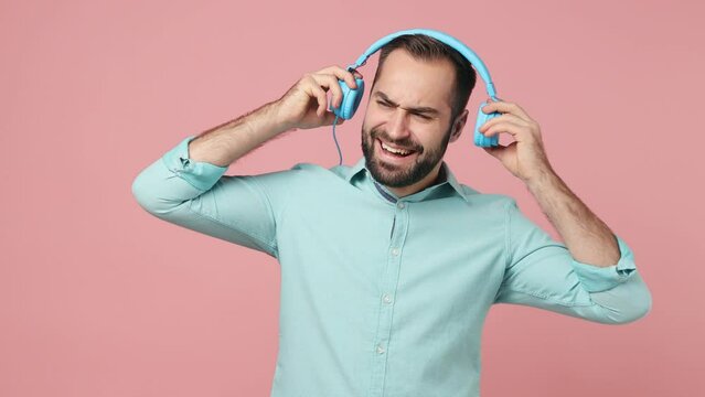 Cheerful vivid young brunet man 20s wears blue shirt listen music in headphones dance sing song fooling around have fun put off gets shocked angry say no isolated on plain plain pink background studio