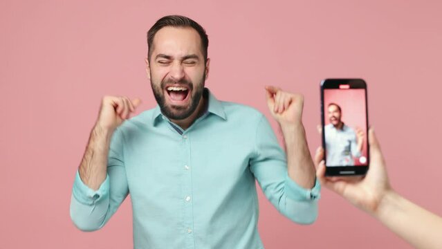 Happy bright cheerful excited fancy young brunet man 20s years old wears blue shirt dancing having fun rest making recording video on mobile cell phone isolated on plain plain pink background studio