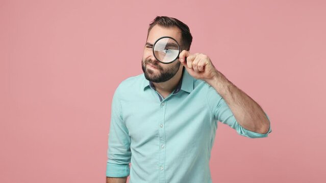 Serious curious businesslike fashionable swanky young bearded brunet man 20s years old wears blue shirt looking camera through magnifying glass isolated on plain plain pink background studio portrait