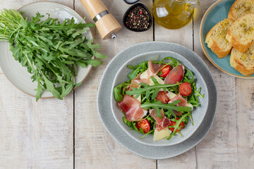 Fresh healthy salad with prosciutto, tomatoes, arugula and cheese in bowl. Baked bread pieces on side. Keto, paleo food. Flat lay