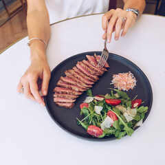 Woman's hand holding fork and eating beef steak with salad, in cafe.