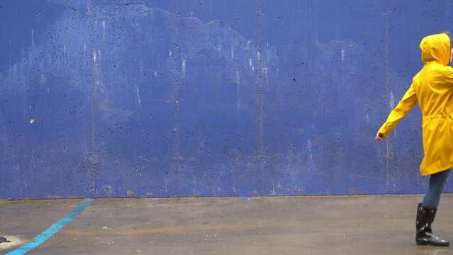 Side view of woman with yellow raincoat walking on blue wall. Full length body of woman under the rain doing big steps with wellingtons and blue background. Seasonal concept winter and spring.