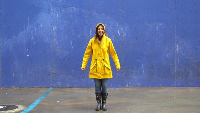 Slow motion front view of woman with yellow raincoat jumping on blue wall. Full length body of woman under the rain leaping with wellington and blue background. Seasonal concept winter and spring.