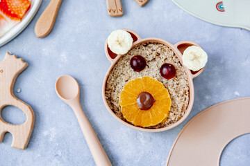 Funny bowl with oat porridge with bear faces made of fruits and berries on blue background. Food for kids idea, top view, copy space.