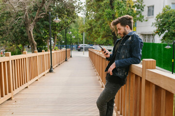 Fototapeta premium Young man looking to mobile phone screen while leaning to wooden fence and wearing denim clothes. Alone man, thinking concepts or watching videos or reading message or scrolling on social media.