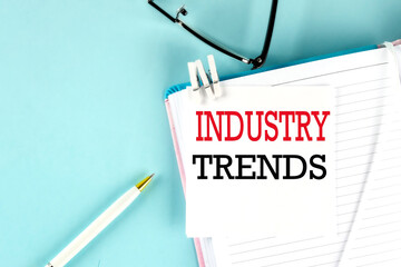 INDUSTRY TRENDS text on a sticky on notebook with pen and glasses , blue background