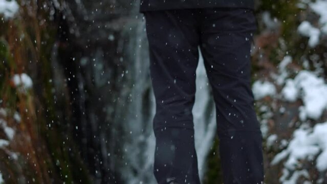 Closeup slow motion shot of snowfall with legs of a man standing in front of Jana waterfall in the background at Kullu in Himachal Pradesh, India